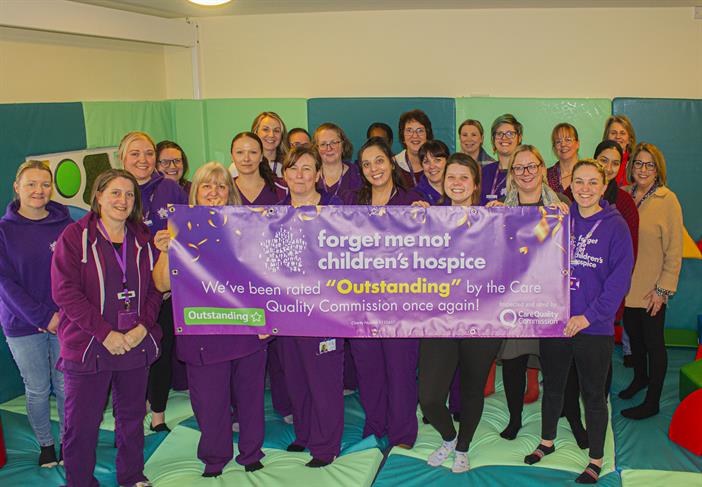 A large group of staff from across forget me not stood in the soft play area, holding a large banner which celebrates the latest outstanding rating from the Quality Care Commission.