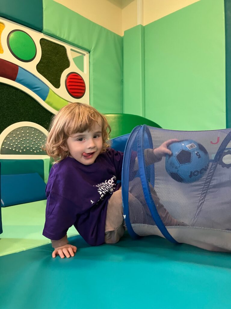 A young white boy with long blonde curly hair emerging from a play tunnel in a soft play area. He is wearing a purple forget me not t-shirt and holding a football in his hand.