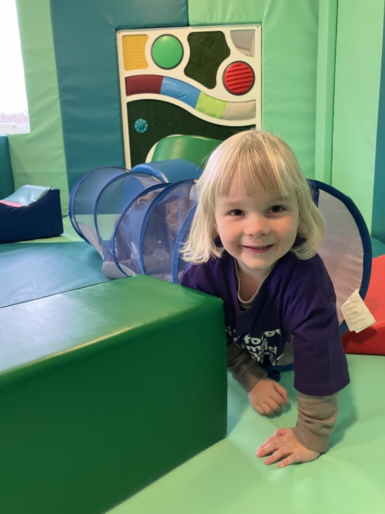 A young white boy with long blonde hair emerging from a play tunnel in a soft play area. He is wearing a purple forget me not t-shirt and smiling at the camera