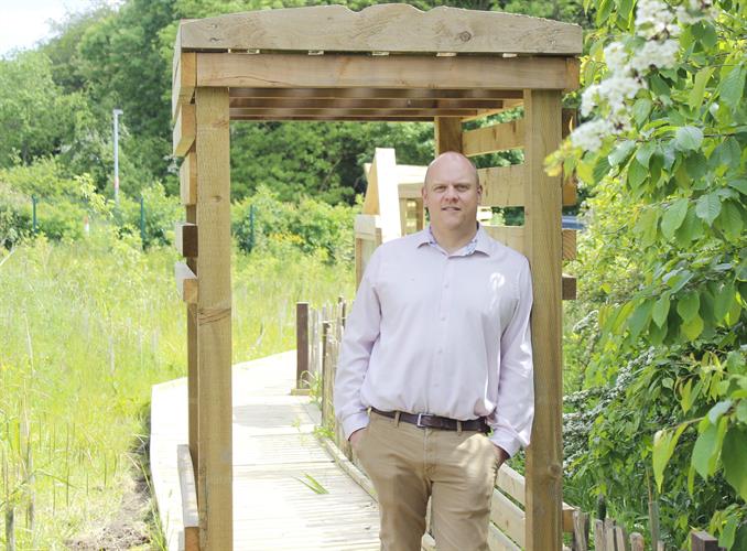 CEO Gareth Pierce stood in a sunny garden at Forget Me Not. He is looking at the camera and leaning against a wooden arch, surrounded by greenery.