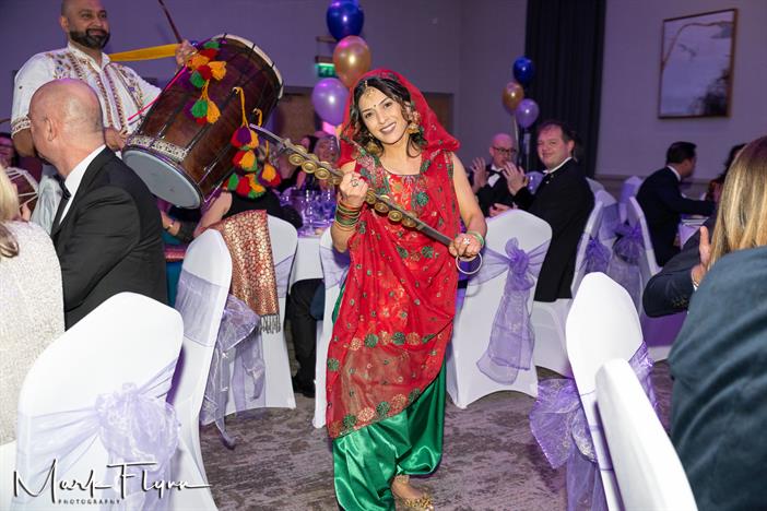 An Asian female wearing red and green traditional dress holding a musical instrument at a dinner event. There are tables of attendees in the background.