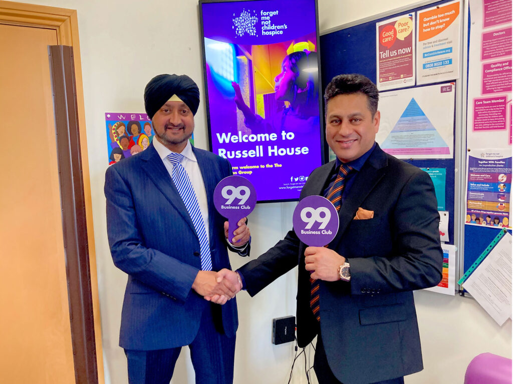 2 Asian business men shaking hands and each holding a 99er business club sign, stood in front of a welcome sign at Russell House