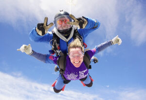 A forget me not fundraiser in the sky, doing a tandem skydive with an instructor on their back. They are both smiling at the camera.