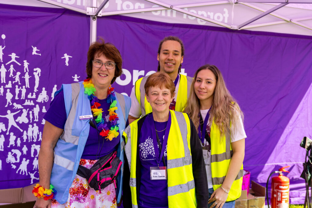 A female member of forget me not staff with 3 volunteers, all wearing high visibility vests. They are stood together at the colour run event and all smiling at the camera.