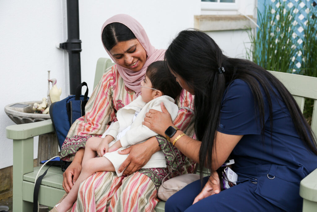 An Asian woman sits with her son on her knee and smiles at him, while an Asian member of the family support team leans in to speak to the boy who has a breathing tube,