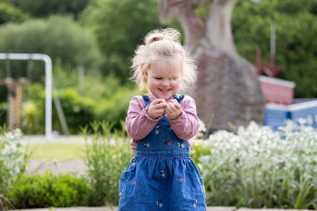 A young white girl with blonde hair is stood in the garden of Russell House, looking and smiling at rocks she's holding in her hand.