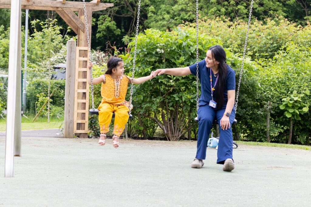 An Asian member of the family support team sits with a young Asian girl on some swings in the garden at Russell House. They are reaching out holding hands and smiling at each other.