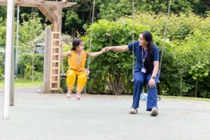 An Asian member of the family support team sits with a young Asian girl on some swings in the garden at Russell House. They are reaching out holding hands and smiling at each other.