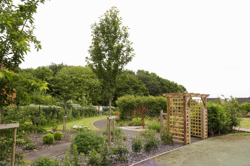 The memory garden showing a small wooden archway leading to a path that leads past the metal memory tree sculpture and beyond.