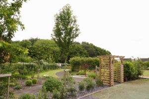 The memory garden showing a small wooden archway leading to a path that leads past the metal memory tree sculpture and beyond.