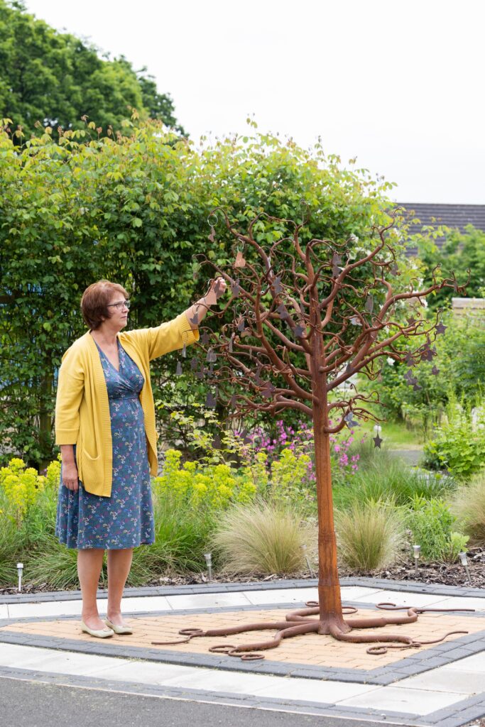 A Forget Me Not employee stoof next to the metal memory tree sculpture in the garden at Russell House, reaching up to read one of the names.