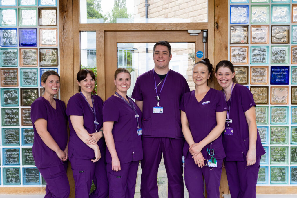 A group from the Forget Me Not care team. A male in the middle, with 5 female staff members stood to the sides, all wearing purple scrubs in front of the coloured glass blocks in the memory corridor at Russell House.