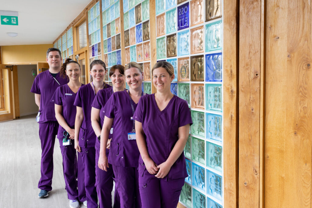 A group from the Forget Me Not care team. A male at the back, with 5 female staff members in a row, all wearing purple scrubs, next to the coloured glass blocks in the memory corridor at Russell House.