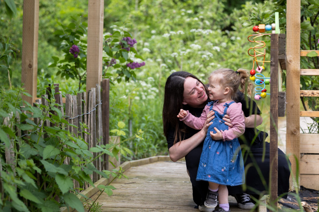 A mum kneels down and holds her young daughter, smiling at her, as they are stood in the garden at Russell House.