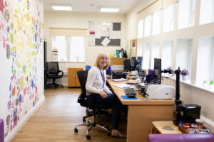 A female volunteer receptionist with blonde hair smiles as she sits behind her desk in the entrance of Russell House.