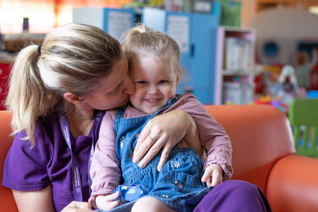 A member of the care team sits in the Hub at Russell House with a young blonde haired girl on her knee. She is holding the girl and kissing her on the cheek while the girl smiles at the camera.