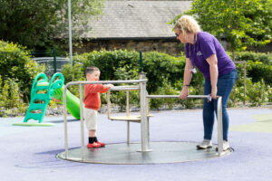 A white female member of staff playing on a roundabout in the outdoor play area of Russell House with a young white boy