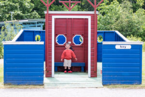 A young white boy with brown hair stood inside a large red and blue wooden boat sculpture outside in a garden setting