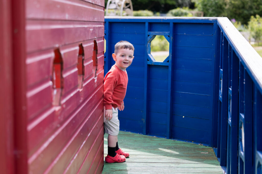 A small white boy with brown hair peering at the camera from behind a wooden play area in the gardens of Russell House.