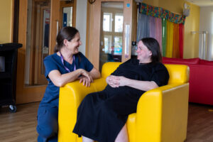 In the hub at Russell House, a white female member of the family support team is knelt down beside a chair where a woman with brown hair and a black dress is sitting. They are smiling at each other.