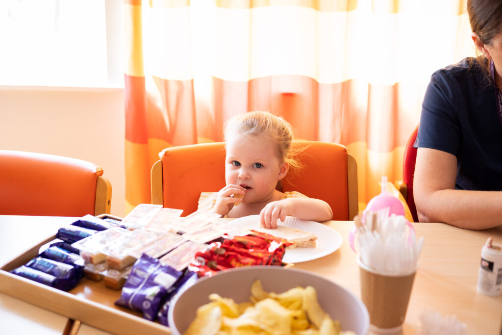 A young while girl with blonde hair sat at a table and eating a sandwich from a plate