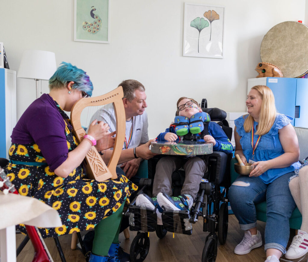 Inside a music therapy session with a young boy in a wheelchair. He is surrounded by his mum and dad, while the music therapist sits beside them playing a harp.