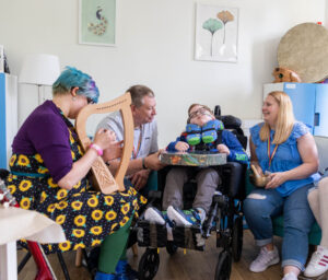 Inside a music therapy session with a young boy in a wheelchair. He is surrounded by his mum and dad, while the music therapist sits beside them playing a harp.