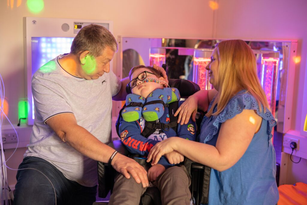 A young white boy in a wheelchair is surrounded by his mum and dad, who are both holding and smiling at him, in the sensory play room at Russell House.