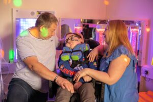 A young white boy in a wheelchair is surrounded by his mum and dad, who are both holding and smiling at him, in the sensory play room at Russell House.
