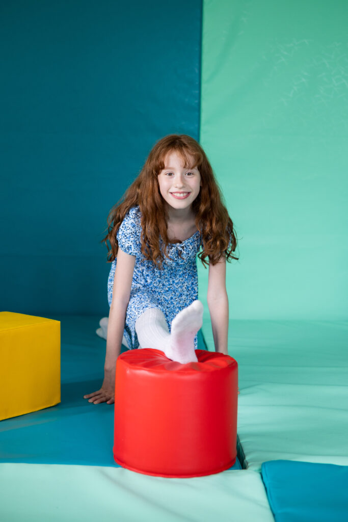 A white girl with long red hair looks at the camera and smiles as she stretches over the play equipment in the soft play room at Russell House