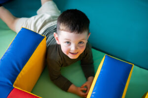 A young white boy with brown hair is lay on the floor of the soft play room next to some soft play equipment, looking up at the camera.