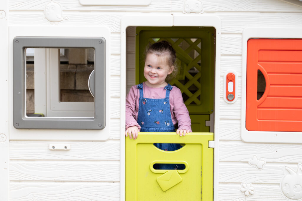 A young white girl with blonde hair is stood at the door of an outdoor play house, looking out and smiling.