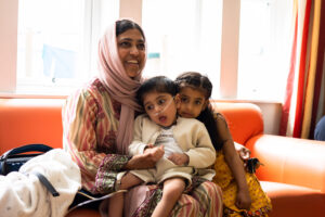 An Asian mum sits in the hub at Russell House while her son, who has a breathing tubs, sits on her lap and her young daughter sits beside them on the sofa.