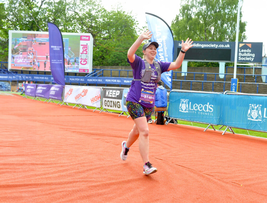 A female competitor finishing the Leeds Marathon, wearing a purple forget me not t-shirt and raising her arms across the finish line
