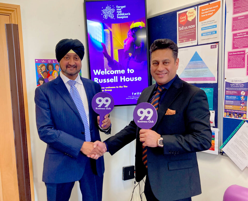 2 Asian business men shaking hands and each holding a 99er business club sign, stood in front of a welcome sign at Russell House