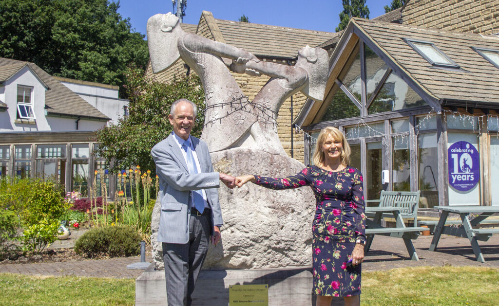 Alison stands in front of the tall stone sculpture in the garden of Russell House and fist bumps the hand of a man beside her as they both smile at the camera.