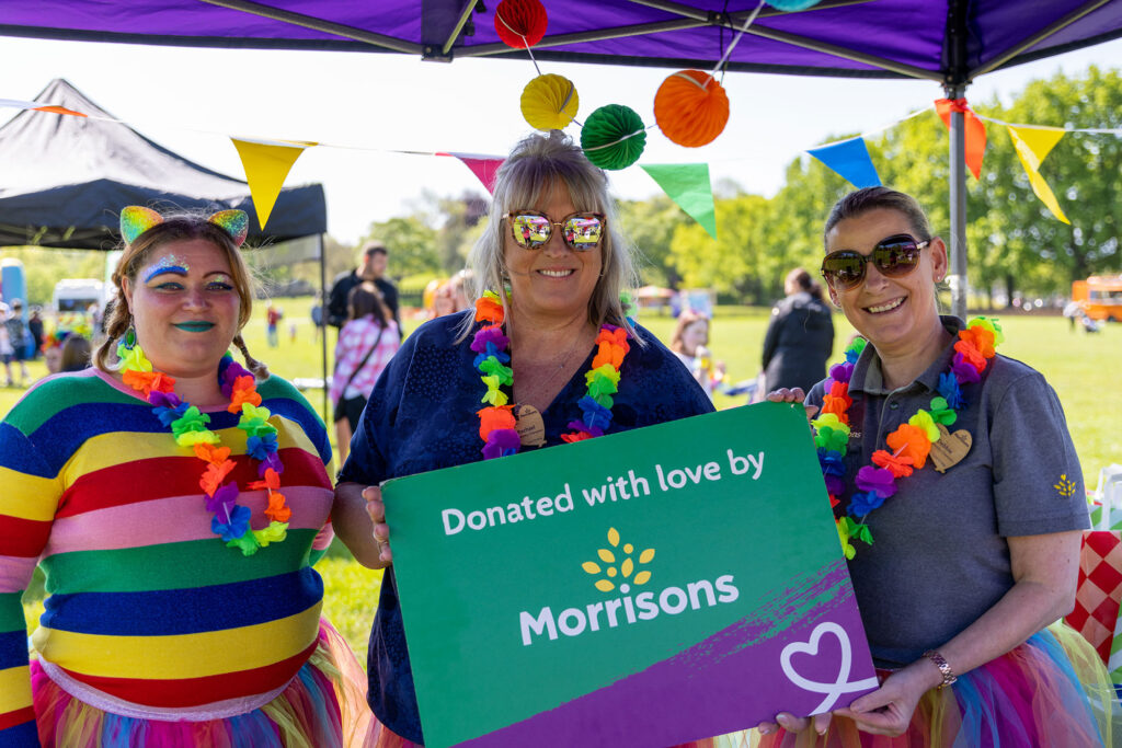 Three women at an outdoor event, wearing bright rainbow coloured leis and tutus, holding a sign that says donated with love by Morrisons