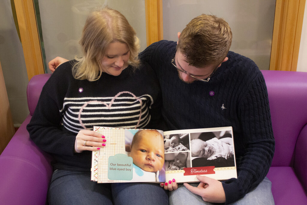 Two parents sit holding open a memory book of their son who they have lost. They are holding the book open and looking down at the pages.