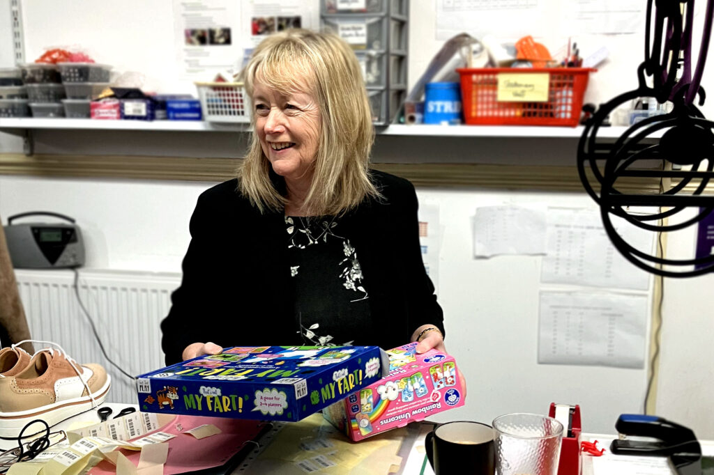 A white female volunteer with blonde hair works in the donation sorting section of the Holmfirth shop. She is holding two board games reading for tagging.