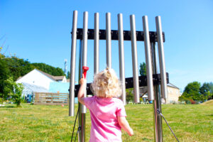 The back of a young girl with curly blonde hair and a pink t-shirt as she hits a musical instrument made of large metal pipes with a beater in the garden. Russell House is in the background.