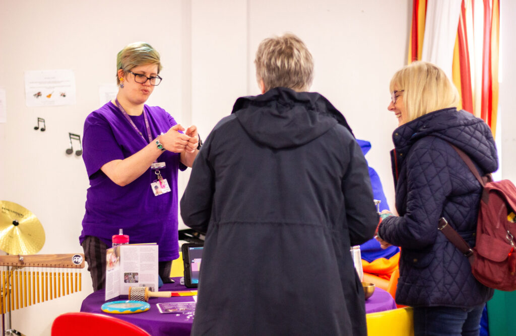 Music Therapist Alice stood speaking to two female visitors on an open day, with a collection of instruments and flyers on the table between them.