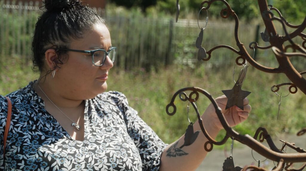 A mum with dark hair and glasses stands in the memory garden at Russell House. She is stood looking at the metal memory tree sculpture and holding a metal heart charm that hangs from it in her hand to read the name on it.