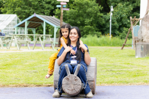An Asian member of the family support team sat on a wooden train sculpture in the garden, with a young Asian girl sat behind her with her arms around her shoulders. They are both smiling at the camera.