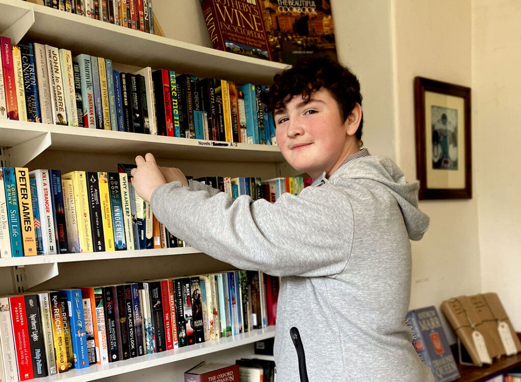 A young white male volunteer with brown curly hair, wearing a light grey tracksuit, is sorting books on a bookshelf at the Holmfirth shop.