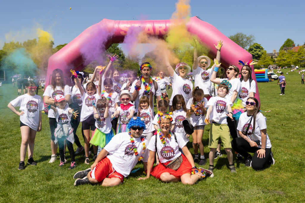 A large group of adults and children having a group photo taken at starting line of the Colour Run event. They are all wearing colour run t-shirts and some are throwing coloured paint powder into the air.