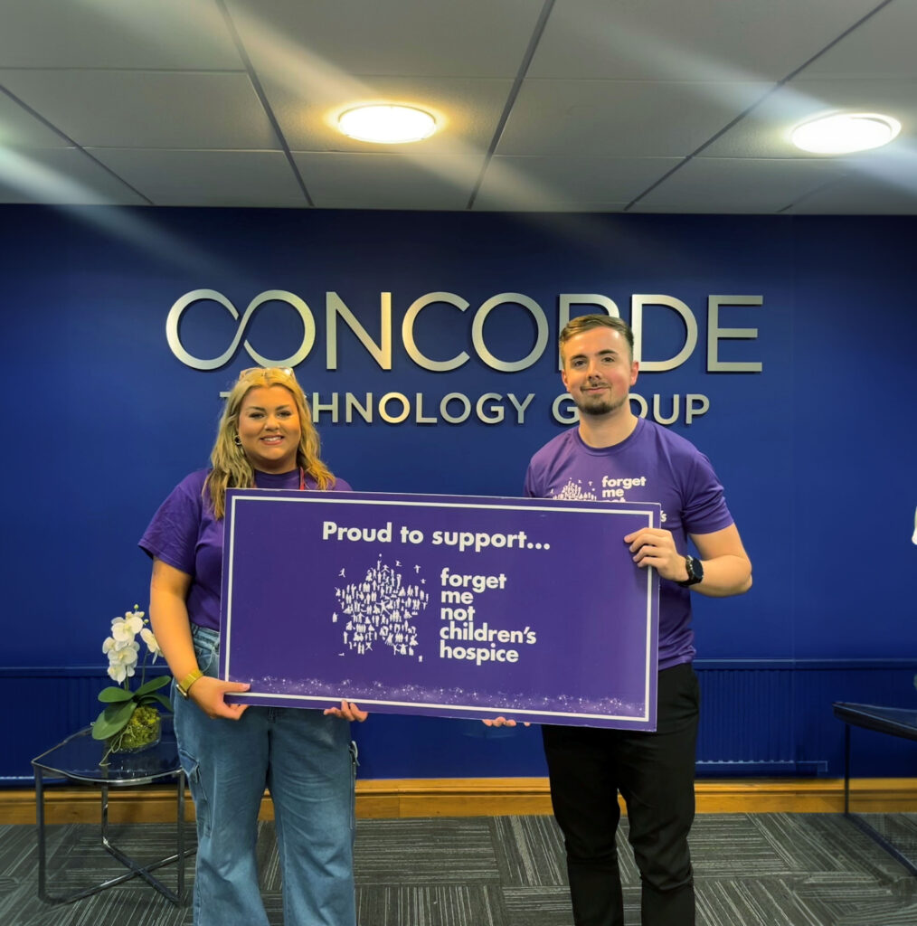 Fundraiser Alex holds a large sign saying "proud to support Forget Me Not Children's Hospice" with a male representative from the Concorde group. They are both wearing Forget Me Not t-shirts.