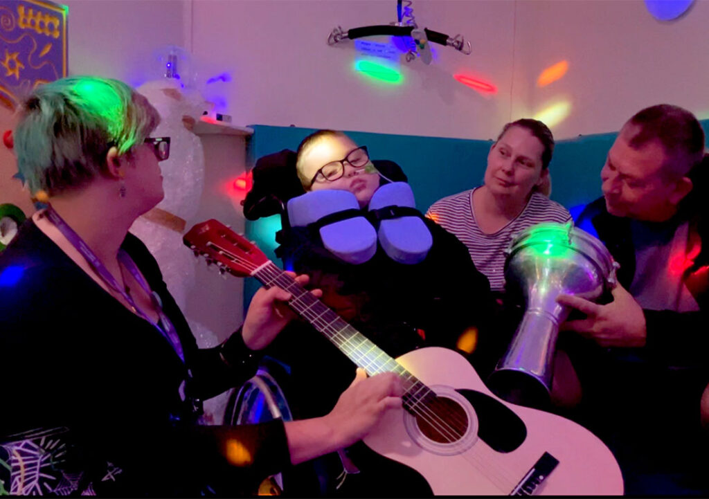 A boy sits in his wheelchair in the sensory room at Russell House, with calming coloured spot lighting across the image. He is surrounded by him parents and the music therapist, who are all holding musical instruments.