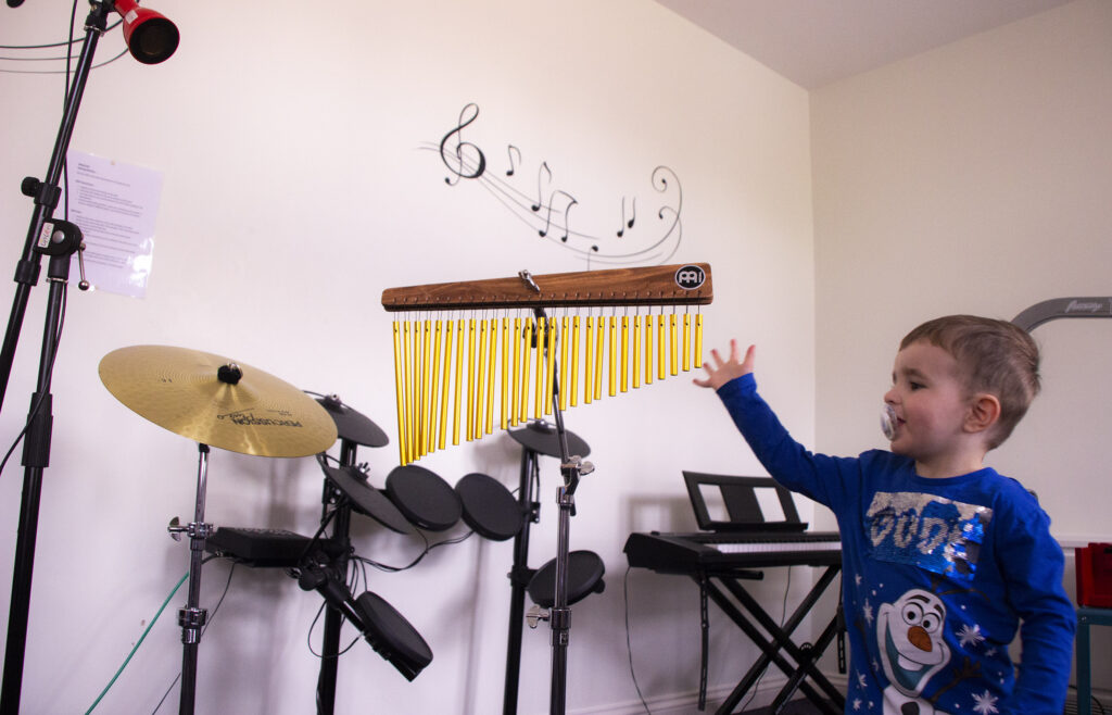 A small boy in the music therapy room, surrounded by different instruments, as he reaches out towards a set of metal chimes.