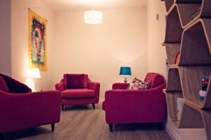 The counselling room at Russell House, showing large comfy red chairs and a sofa, a colourful tapestry on the wall and a set of shelves holding toys and books.
