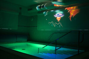 The hydrotherapy pool at Russell House in low light, with the pool illuminated green and blue by underwater lighting and space-themed images projected onto the wall.
